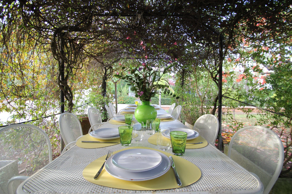 Dining table set for four with green glasses and plates, surrounded by white chairs under a floral arbor.